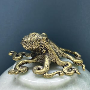 May include: A small, textured brass octopus figurine with eight curved tentacles, featuring a detailed, bumpy surface on its head and body, resting atop a smooth, pale green, rounded stone base with a polished finish and subtle marbling.