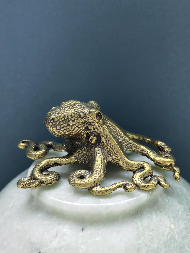 May include: A small, textured brass octopus figurine with eight curved tentacles, featuring a detailed, bumpy surface on its head and body, resting atop a smooth, pale green, rounded stone base with a polished finish and subtle marbling.