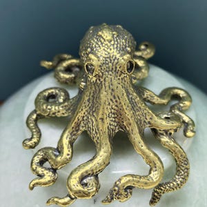 May include: A brass-colored metal octopus figurine featuring a textured surface, a rounded head, and eight curved, sprawling tentacles. The octopus has a metallic finish with dark recessed areas that highlight the detailed skin texture.