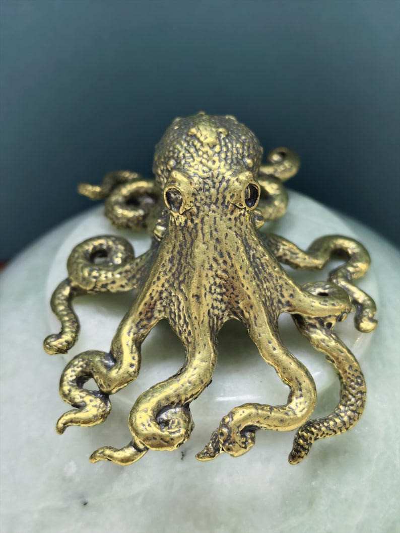 May include: A brass-colored metal octopus figurine featuring a textured surface, a rounded head, and eight curved, sprawling tentacles. The octopus has a metallic finish with dark recessed areas that highlight the detailed skin texture.