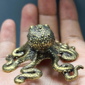 May include: A small, three-dimensional brass octopus figurine with a textured, bumpy surface on its rounded head and eight curved, detailed tentacles extending outward. The metal has a dark, antiqued gold finish with visible highlights.