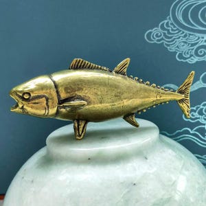 May include: A metallic gold-toned tuna fish figurine with detailed fins and scales, positioned horizontally. The fish has an open mouth and is resting on top of a smooth, rounded, pale green stone object with a polished, mottled surface texture.