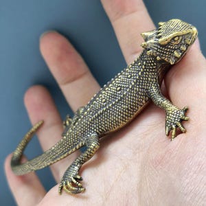 Puede incluir: Una pequeña figura de lagarto de metal en tono dorado con una superficie texturizada y escamosa. El lagarto presenta una cabeza detallada con púas prominentes, una cola larga y cuatro patas con garras distintas, con una pátina oscura.