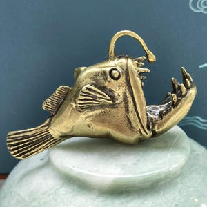 Peut inclure: Une figurine en métal couleur laiton représentant une baudroie avec un leurre proéminent sur la tête, une large bouche ouverte garnie de dents pointues, des nageoires texturées et une queue détaillée, vue de profil avec une finition métallique.