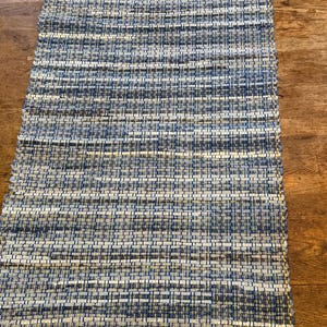 May include: A rectangular woven runner rug featuring a textured pattern of horizontal stripes in shades of navy blue, light blue, white, and grey. The rug is finished with a decorative fringe along the bottom edge in a solid golden yellow color.