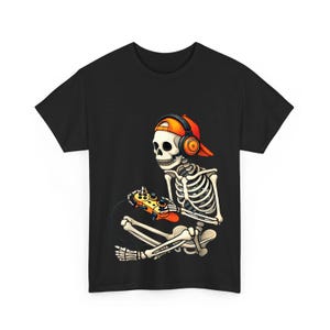 May include: A black t-shirt featuring a graphic of a white skeleton wearing an orange baseball cap and black headphones. The skeleton is sitting down and holding an orange video game controller with both hands, focused on playing a game.