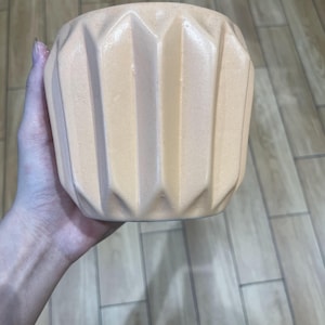 Könnte beinhalten: Ein pfirsichfarbener Keramikübertopf mit einer Reihe vertikaler, kantiger Rippen, die eine geometrische, facettierte Textur an der Außenseite erzeugen. Der Topf hat eine breite, offene Oberseite und eine leicht konisch zulaufende Basis.