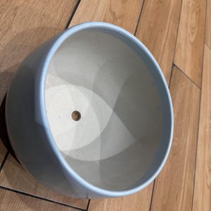 Könnte beinhalten: Ein runder, hellblauer Keramik-Pflanztopf mit einer glatten, glänzenden Außenfläche. Das Innere ist matt cremeweiß und verfügt über ein einzelnes, mittig platziertes Abflussloch am Boden des Beckens, das für die Pflanzenpflege vorgesehen ist.