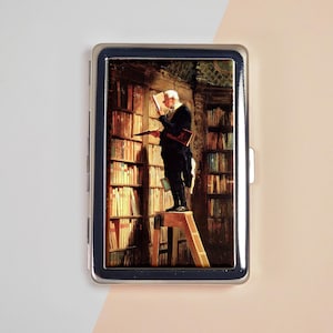 May include: A rectangular metal case with a silver-toned frame featuring a printed image of a person standing on a wooden step ladder in a library, reading a book while surrounded by tall, dark wooden bookshelves filled with numerous books.