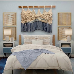 Puede incluir: Un gran tapiz de pared de fibra con nudos texturizados de color crema y flecos azul oscuro suspendido de una barra de madera. La pieza se exhibe sobre una cama con un edredón blanco estampado, una manta azul y dos mesitas de noche.