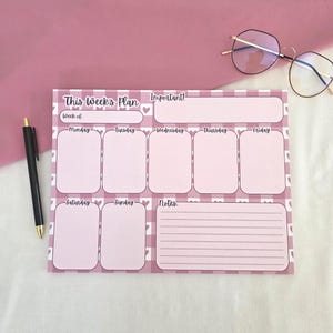 May include: A rectangular weekly planner notepad with a pink and white checkered pattern featuring small hearts. The layout includes sections labeled This Week's Plan, Important, days of the week from Monday to Sunday, and a lined Notes section.