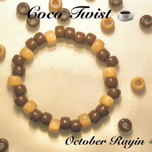 Può includere: Un braccialetto circolare composto da perline cilindriche in plastica che si alternano tra il marrone scuro e il beige chiaro. L'immagine include il testo Coco Twist e October Rayin, insieme a una piccola grafica di una tazza di caffè e un'icona di fiore di loto rosa.