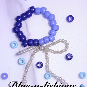 Puede incluir: Una pulsera circular hecha de cuentas de plástico azul oscuro y azul claro, con un dije de lazo colgante hecho de pequeñas perlas blancas. El texto Blue-a-lishious está impreso en negro en la parte inferior, rodeado de cuentas azules sueltas.