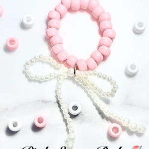Puede incluir: Una pulsera hecha de cuentas de plástico de color rosa claro con un lazo decorativo formado por pequeñas perlas blancas esféricas. El texto October Rayin y Pink Sugar Baby aparece impreso en la imagen junto a cuentas sueltas dispersas.