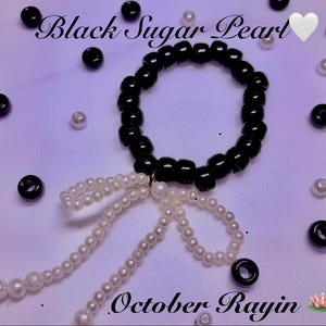 Puede incluir: Una pulsera de cuentas negras brillantes con un dije colgante de lazo hecho de perlas blancas. El texto Black Sugar Pearl y October Rayin es visible. Cuentas negras y blancas dispersas rodean la pulsera central sobre un fondo morado claro.