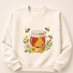 Könnte beinhalten: Ein cremefarbenes Sweatshirt mit Rundhalsausschnitt und einer Stickerei, die ein Glas mit bernsteinfarbenem Honig zeigt. Das Glas ist mit einem Wabenmuster, einer gebundenen Schnur, drei fliegenden Bienen, einem Honiglöffel und gelben Blumen verziert.