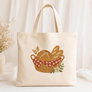 Puede incluir: Un bolso de tela color crema con un diseño bordado de una cesta de mimbre llena de panes. La cesta está forrada con un paño de cuadros rojos y blancos y decorada con espigas de trigo y una pequeña flor blanca.