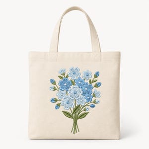 May include: A natural-colored canvas tote bag featuring a centered machine embroidery design of a bouquet of blue flowers. The floral arrangement includes various shades of light and medium blue petals, green stems, and small green leaves.
