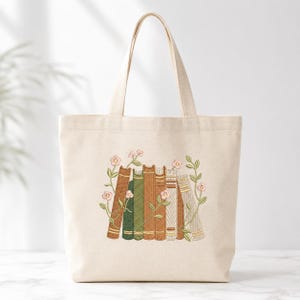 May include: A cream-colored canvas tote bag featuring an embroidered design of a row of upright books in shades of brown, green, and beige. The books are intertwined with delicate green vines and small pink flowers, creating a detailed illustration.