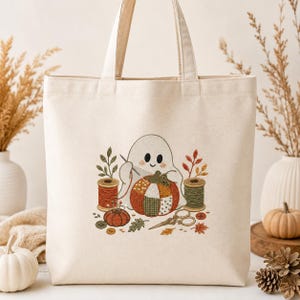 May include: A cream-coloured canvas tote bag featuring a central graphic of a white ghost sewing a patchwork pumpkin. The design includes two spools of thread, a pincushion, scissors, buttons, and autumn leaves in shades of orange, green, and brown.