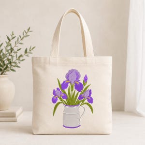 May include: A cream-colored canvas tote bag featuring a centered graphic illustration of purple iris flowers arranged in a light gray pitcher. The design includes green stems and leaves, with the pitcher outlined in a thin purple border.