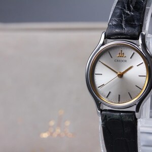 May include: A Credor wristwatch featuring a round silver dial with gold-tone hands and hour markers, encased in a polished silver-tone metal frame with a gold inner ring. The watch is attached to a black textured leather strap with a visible grain.