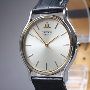 May include: A Credor Seiko wristwatch with a round silver dial, gold-tone hands, and thin gold-tone hour markers. The watch features a silver-tone bezel with a thin gold-tone inner ring, a black leather strap, and the Credor logo at the twelve o'clock position.