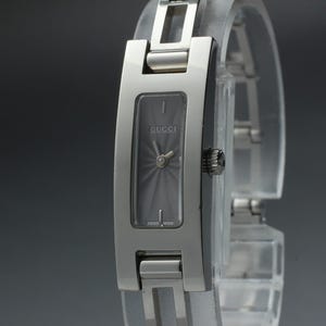 May include: A silver-toned stainless steel Gucci wristwatch featuring a rectangular case with a dark grey sunburst dial. The watch has a matching metal link bracelet and a small crown on the right side. The dial displays the Gucci logo and Swiss Made text.