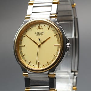 May include: A Credor Seiko wristwatch featuring a round, pale yellow dial with gold-tone hour markers and hands. The watch has a two-tone stainless steel and gold-tone metal link bracelet. The Credor logo is positioned at the twelve o'clock mark.