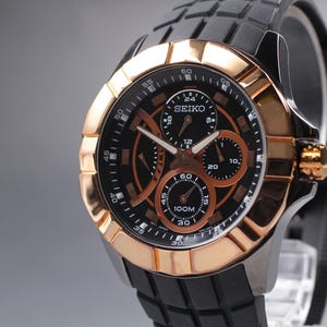 May include: A Seiko chronograph watch featuring a black dial with rose gold-tone accents, three sub-dials, and a date display. The watch has a rose gold-tone bezel and a black textured silicone strap with a matching black case and crown.