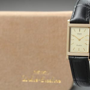 May include: A rectangular Seiko Quartz Exalina wristwatch featuring a gold-tone case, a cream-colored dial with thin gold hour markers, and a black leather strap with a textured finish. The watch face displays the Seiko Quartz and Exalina branding.