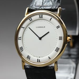 May include: A Lassale wristwatch with a round gold-tone case and a white dial featuring black Roman numerals. The watch has thin black hands and a textured black leather strap. The word Lassale is printed in black on the center of the dial.