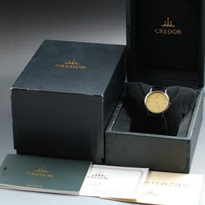 May include: A Credor wristwatch with a round gold-toned face and a black leather strap, presented in a black hinged box with the Credor logo printed in gold. Included are two small booklets with the Credor logo and Japanese text on the covers.