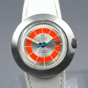 Pode incluir: Um relógio automático Omega Genève Dynamic com caixa redonda de aço inoxidável e bracelete de couro branco. O mostrador apresenta um centro prateado, um anel circular laranja brilhante, ponteiros brancos e um ponteiro de segundos azul.
