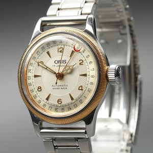 May include: An Oris automatic wristwatch with a silver-tone stainless steel link bracelet and a round case featuring a gold-tone fluted bezel. The cream-colored dial displays Arabic numerals, a date pointer hand with a red crescent, and text.