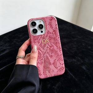 Puede incluir: Una funda para smartphone de color rosa y blanco con un patr&oacute;n texturizado de piel de serpiente y las letras doradas QS grabadas en el centro. La funda presenta un recorte rectangular para un sistema de c&aacute;mara de tres lentes en la esquina superior izquierda.