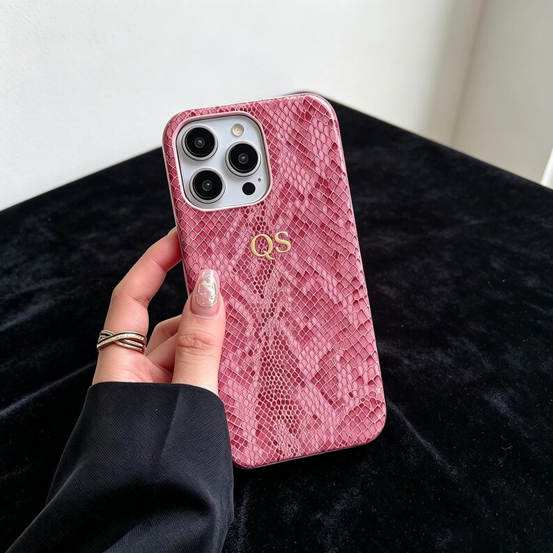 Puede incluir: Una funda para smartphone de color rosa y blanco con un patr&oacute;n texturizado de piel de serpiente y las letras doradas QS grabadas en el centro. La funda presenta un recorte rectangular para un sistema de c&aacute;mara de tres lentes en la esquina superior izquierda.