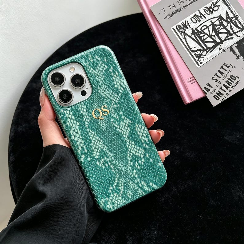 Puede incluir: Una funda para tel&eacute;fono con estampado de piel de serpiente en verde y blanco, con un recorte para triple c&aacute;mara y las iniciales doradas QS centradas en la parte posterior. La funda tiene una superficie texturizada para un smartphone de tres lentes.