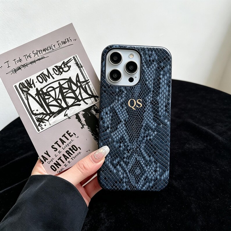 Puede incluir: Una funda de tel&eacute;fono con estampado de piel de serpiente en azul oscuro y negro, con las iniciales QS grabadas en oro en el centro. La funda tiene un recorte para un sistema de triple c&aacute;mara y se sostiene junto a una tarjeta con texto graffiti.