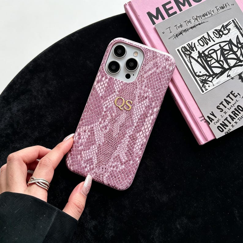 Puede incluir: Una funda de smartphone rosa y blanca con un patr&oacute;n texturizado de piel de serpiente y las iniciales doradas QS centradas en la parte posterior. La funda cuenta con un recorte para un sistema de triple c&aacute;mara y est&aacute; junto a un libro rosa con texto.