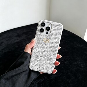 Puede incluir: Una funda de smartphone blanca con un patr&oacute;n texturizado de piel de serpiente y las letras doradas QS grabadas en el centro. La funda cuenta con un recorte para un sistema de c&aacute;mara de triple lente y un flash, protegiendo el dispositivo.