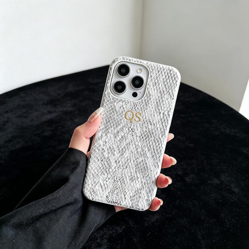 Puede incluir: Una funda de smartphone blanca con un patr&oacute;n texturizado de piel de serpiente y las letras doradas QS grabadas en el centro. La funda cuenta con un recorte para un sistema de c&aacute;mara de triple lente y un flash, protegiendo el dispositivo.