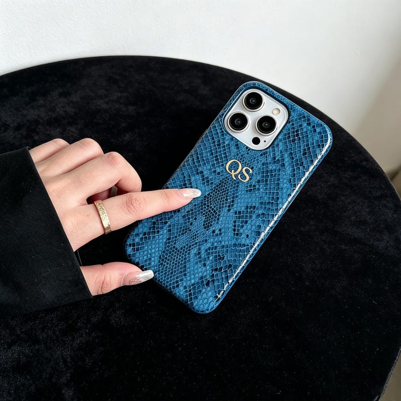 Puede incluir: Una funda de smartphone azul con un patr&oacute;n de piel de serpiente texturizado y las letras doradas QS grabadas en la parte posterior. La funda incluye un recorte para un sistema de c&aacute;mara de triple lente para un dispositivo m&oacute;vil moderno.