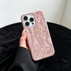 May include: A light pink smartphone case with a textured snakeskin pattern and the gold-colored letters QS embossed in the center. The case features a cutout for a triple-lens camera system and a flash on the upper left corner of the back panel.