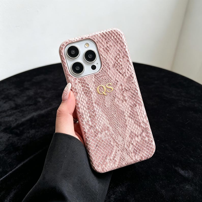 Puede incluir: Una funda para smartphone de color rosa claro con un patr&oacute;n texturizado de piel de serpiente y las letras doradas QS grabadas en el centro. La funda cuenta con un recorte para un sistema de triple c&aacute;mara y flash en la esquina superior.