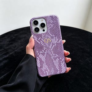 Puede incluir: Una funda de smartphone morada con un patr&oacute;n texturizado de piel de serpiente y las letras doradas QS grabadas en el centro. La funda presenta un recorte para un sistema de triple c&aacute;mara en la esquina superior izquierda, mostrando tres lentes.