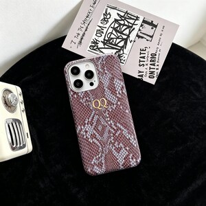 Puede incluir: Una funda para smartphone con un patr&oacute;n de piel de serpiente marr&oacute;n y gris con acabado texturizado. La funda muestra las letras doradas QQ en el centro e incluye un recorte para la c&aacute;mara con tres lentes circulares en el lado izquierdo.