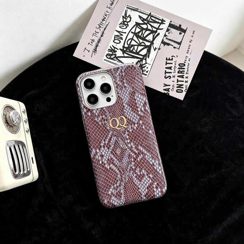 Puede incluir: Una funda para smartphone con un patr&oacute;n de piel de serpiente marr&oacute;n y gris con acabado texturizado. La funda muestra las letras doradas QQ en el centro e incluye un recorte para la c&aacute;mara con tres lentes circulares en el lado izquierdo.