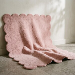 Contemporary pink area rug with a plush cloud-inspired design and distinctive scalloped edges. This decorative floor covering creates a charming accent for bedrooms, nurseries, living rooms, and modern apartments. The soft pink color complements ivory sofas, beige upholstery, light wood furniture, pastel accessories, and simple decorative pieces. Its wavy border provides a playful alternative to conventional rug shapes, making it a stylish choice for cozy Scandinavian and minimalist interiors.