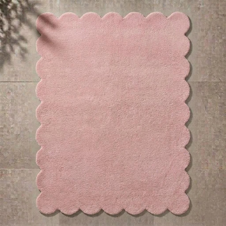 Soft pink cloud rug with a scalloped edge and plush surface, designed for cozy contemporary interiors. This wavy border area carpet brings a gentle decorative touch to nurseries, children's bedrooms, living rooms, and relaxing reading spaces. Pair it with white furniture, pastel cushions, natural wood, soft blankets, and minimalist accessories. The cloud-like silhouette creates visual interest while maintaining a calm, welcoming atmosphere suited to Scandinavian and modern decor.